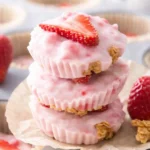 Frozen strawberry yogurt bites with granola base and fresh strawberry topping stacked on parchment paper