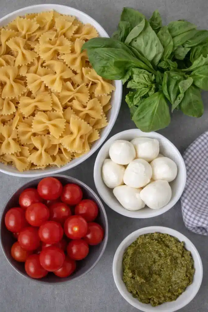 Bow tie pasta salad ingredients including farfalle pasta, basil, mozzarella, tomatoes, and pesto