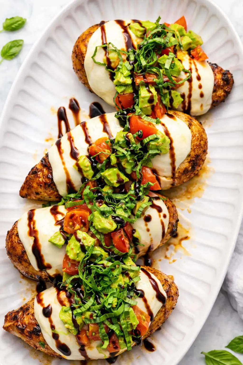 California grilled chicken with mozzarella, avocado, tomatoes, basil and balsamic glaze on a white serving platter.