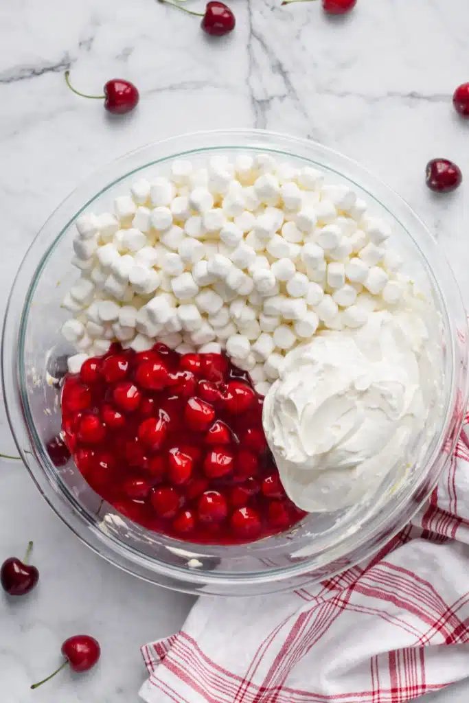 Cherry cheesecake fluff ingredients with marshmallows whipped cream and cherry pie filling in glass bowl