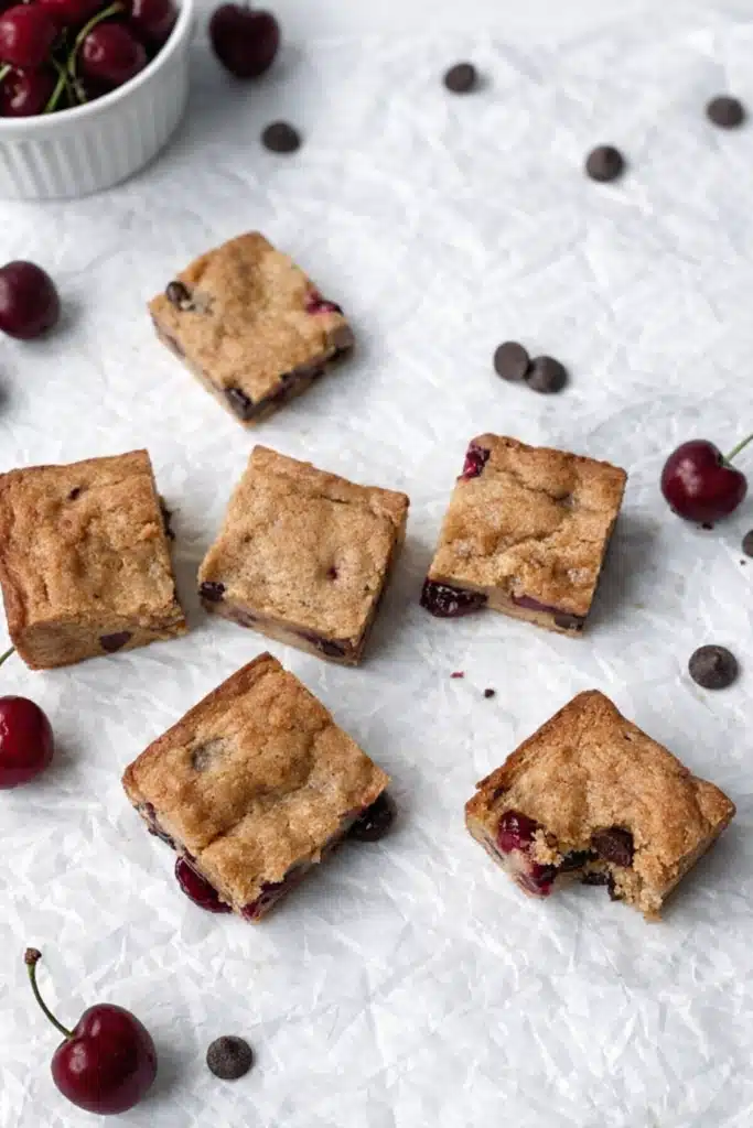 Cherry chocolate chip blondies squares scattered on parchment with fresh cherries