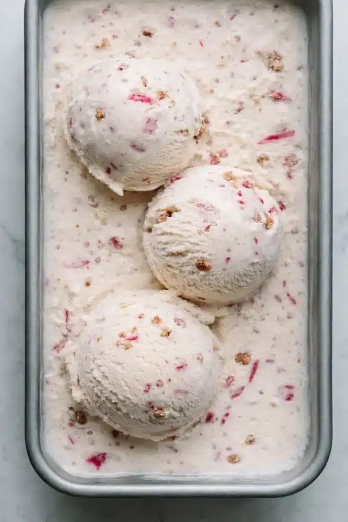 Rhubarb crumble ice cream scoops with pink rhubarb pieces and crunchy crumble in a dessert container