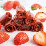 Stacked homemade strawberry fruit roll-ups with fresh strawberries on marble surface