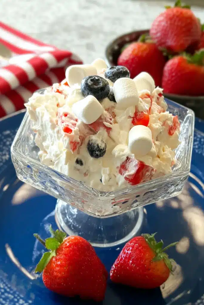 Red White & Blue Cheesecake Salad served in a glass bowl with strawberries blueberries and marshmallows