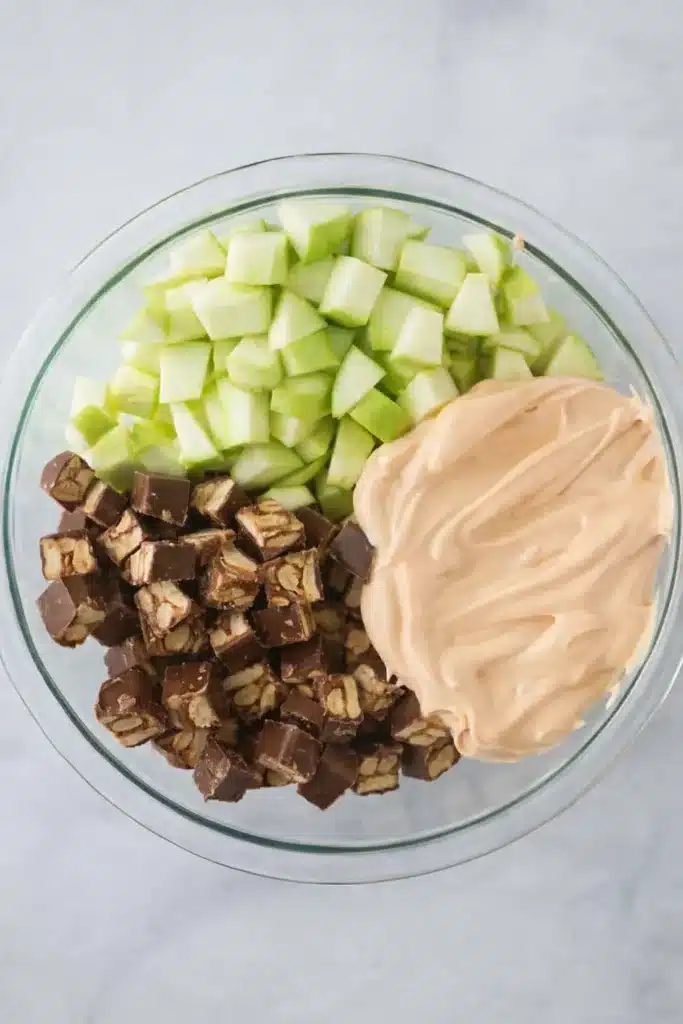 Snickers apple salad ingredients with apples, candy pieces, and creamy pudding in a bowl