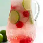 Cherry limeade in a glass pitcher with lime slices, cherries, ice, and sparkling soda