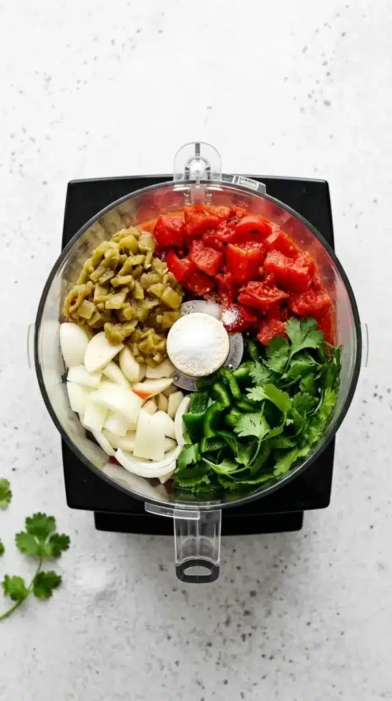 Best Salsa Recipe ingredients in a food processor with tomatoes, cilantro, green chilies, garlic, and jalapeno