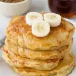 Brown Sugar Banana Pancakes stacked with banana slices and maple syrup on a cream plate