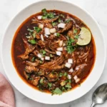 Slow cooker Birria with shredded beef, rich consommé, cilantro, onion, and lime