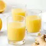 Ginger Shots: Amazing Easy Recipe for a Happy Morning 4 Homemade Ginger Shots in two small glasses with fresh ginger and lemon on a light stone countertop
